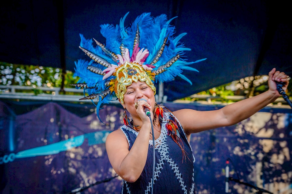 A performer wearing a colorful feathered headdress is passionately singing into a microphone on an outdoor stage, arm raised.