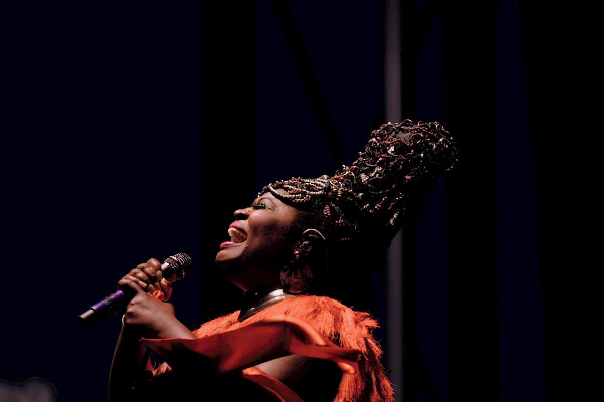 The image shows a person with elaborate hair and attire passionately singing into a microphone against a dark background.