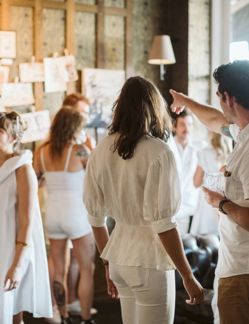 A group of people in white attire are gathered, with one person pointing at a wall of pictures, inside a well-lit room.