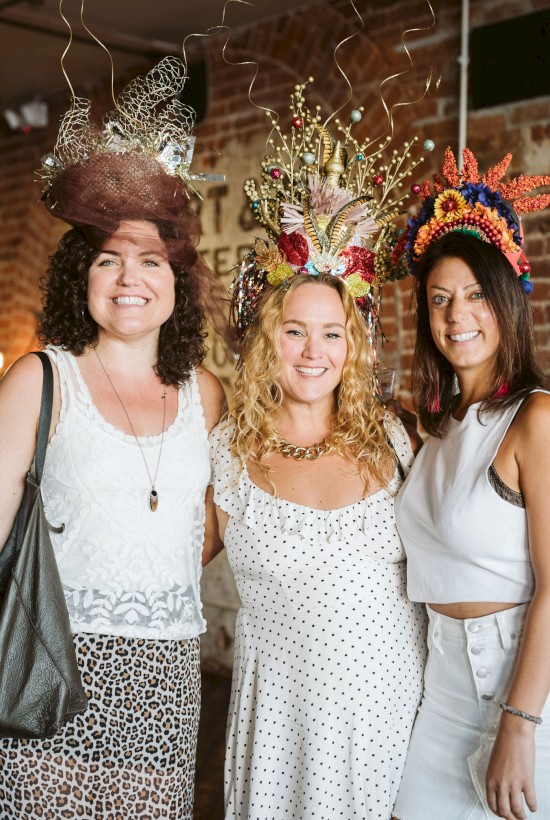 Three people smiling, wearing decorative headpieces, posing indoors with a brick wall and a sign in the background.
