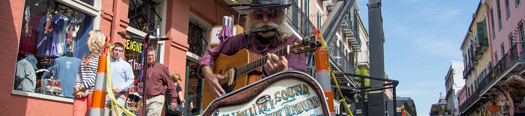 A street performer playing guitar next to an open guitar case with a humorous sign in a lively urban setting.