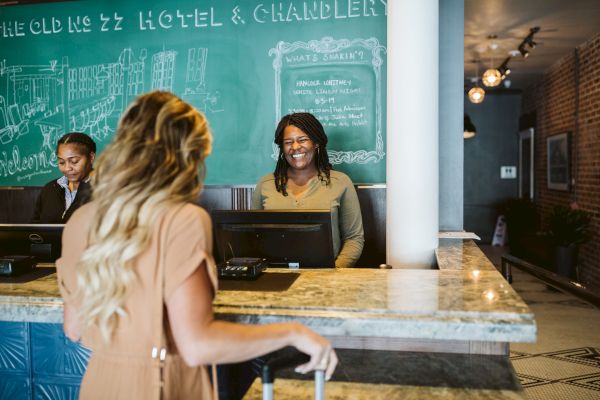 A person is checking in at a hotel reception with two staff members behind the counter, one smiling. There is a chalkboard in the background.