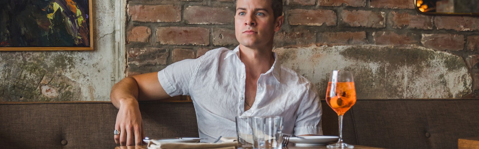 A person sits in a rustic café with exposed brick walls, wearing casual attire. A glass of orange drink is on the table, next to utensils.