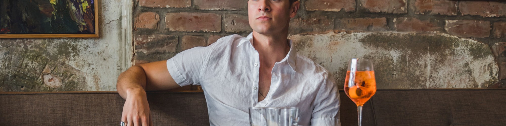 A person sits in a rustic café with exposed brick walls, wearing casual attire. A glass of orange drink is on the table, next to utensils.