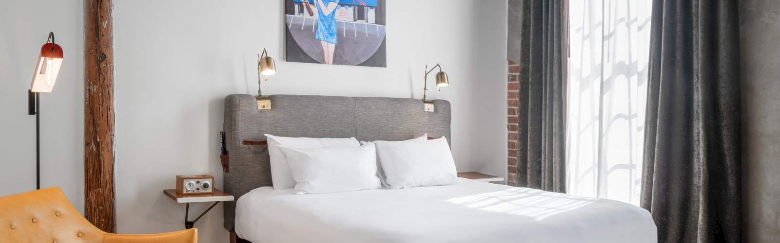 A cozy bedroom with a double bed, exposed brick wall, artwork, desk, TV, and a mix of vintage and modern decor elements.