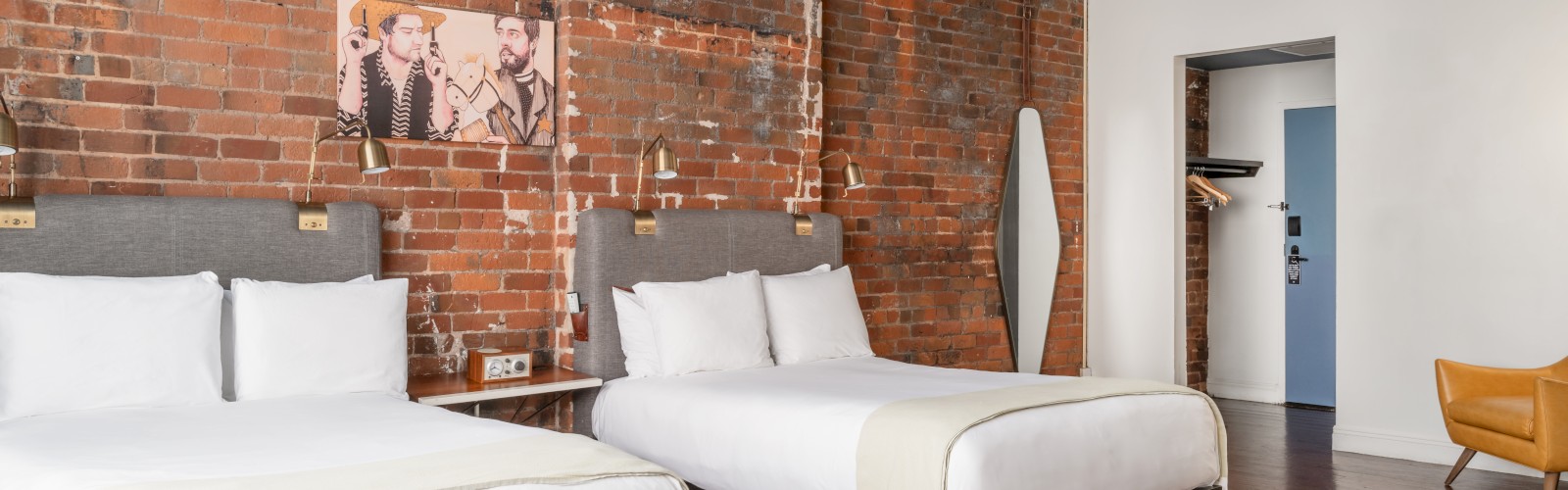 The image shows a cozy hotel room with two double beds, brick walls, stylish modern decor, and a yellow chair by the window.