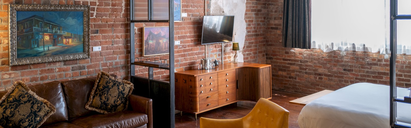 A cozy loft-style room with brick walls, featuring a leather sofa, yellow chairs, wooden furniture, and a neatly made bed by a window.