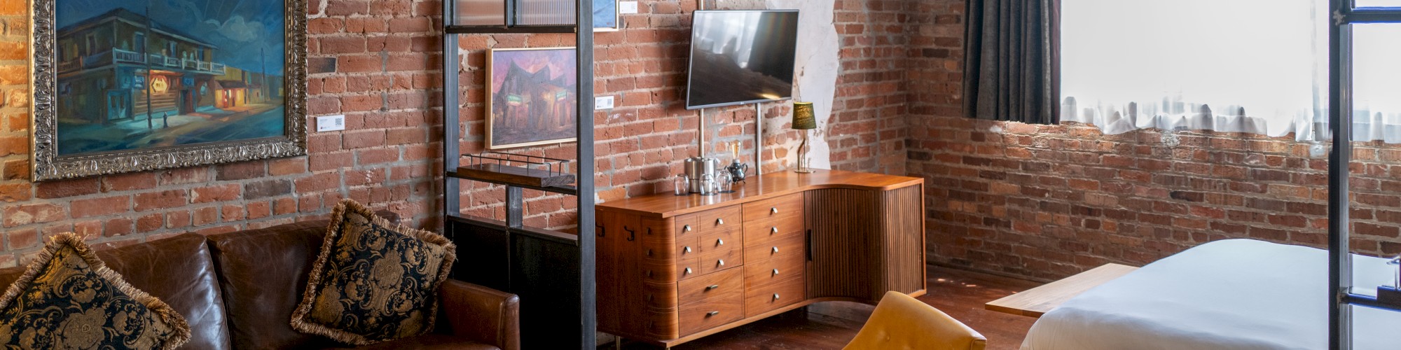 A cozy loft-style room with brick walls, featuring a leather sofa, yellow chairs, wooden furniture, and a neatly made bed by a window.