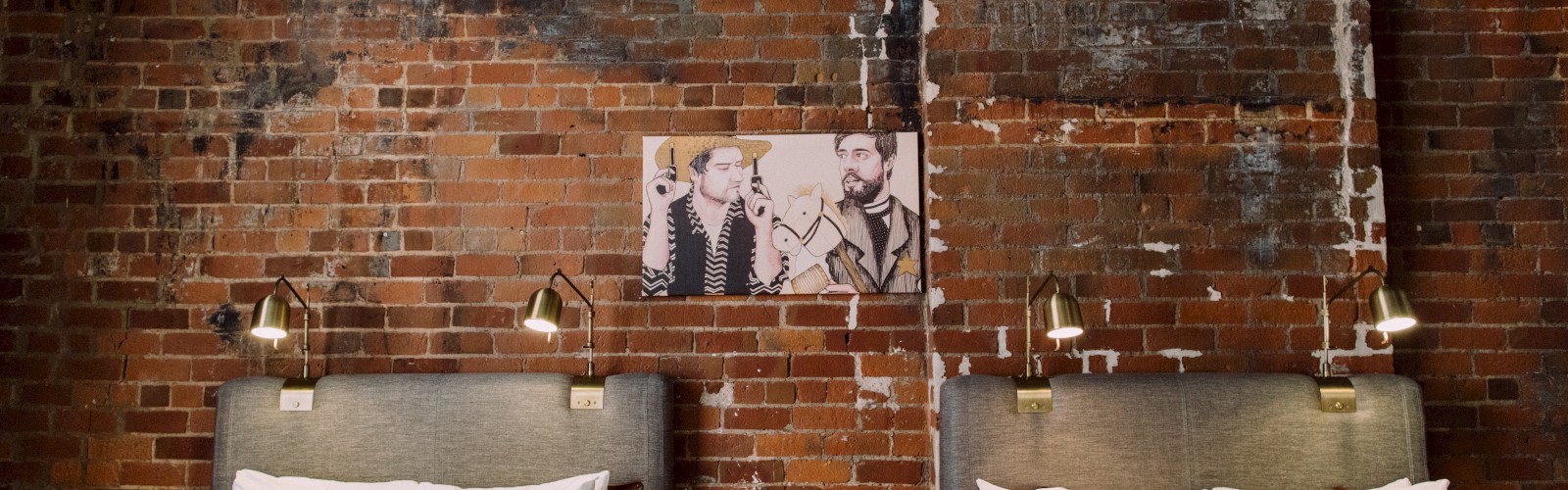 A room with exposed brick walls, two beds with white linens, a central console table, and a picture of three people on the wall above.