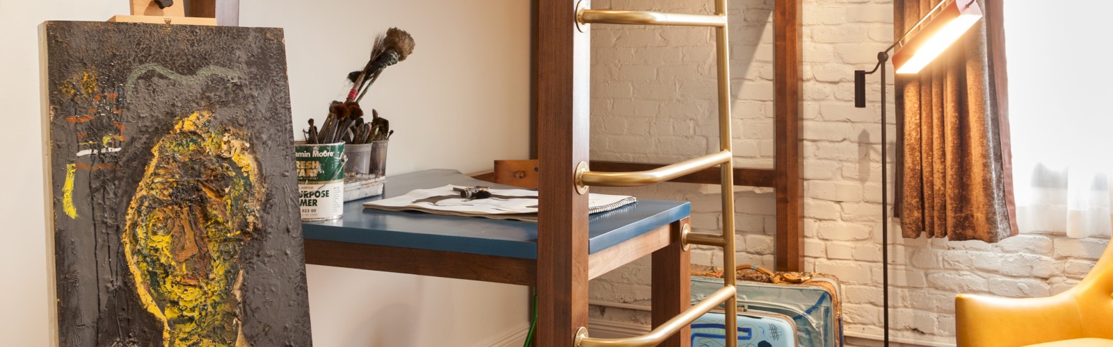 The image shows a cozy room with a loft bed, an easel with a painting, a study desk under the bed, a suitcase, a floor lamp, and a mustard chair.