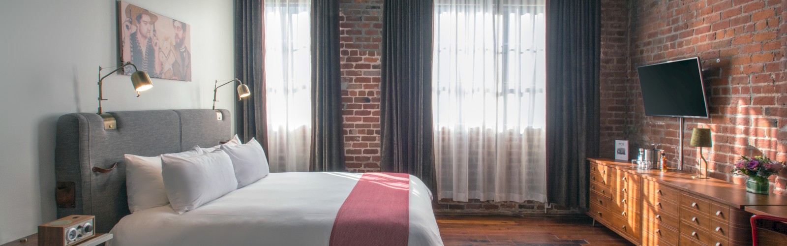 The image shows a cozy bedroom with a brick wall, a bed with white and pink bedding, a TV, and curtains covering large windows.