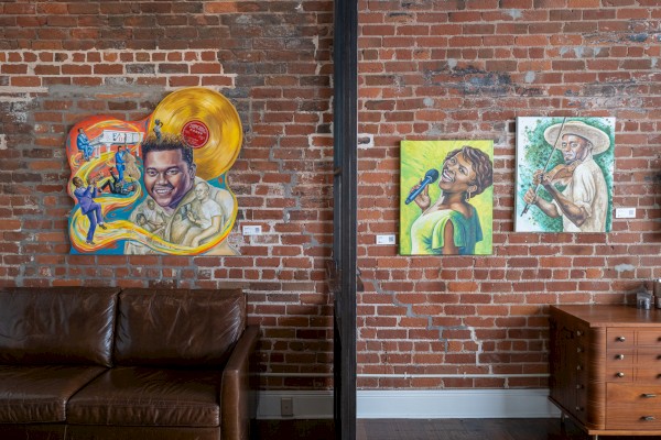 The image shows a brick wall with three colorful paintings of musicians, featuring a couch and a wooden dresser below.