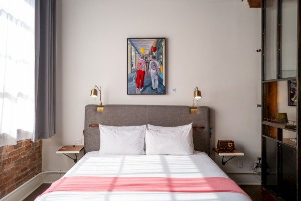 A cozy bedroom with a large bed, decorative pillows, a colorful wall art piece, modern lamps, and natural light streaming through a window.