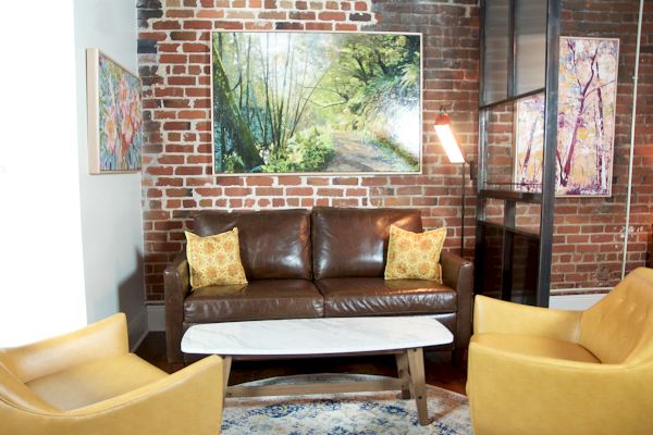 A cozy living room with a brown leather sofa, yellow armchairs, a white coffee table, brick walls, and nature-themed paintings.