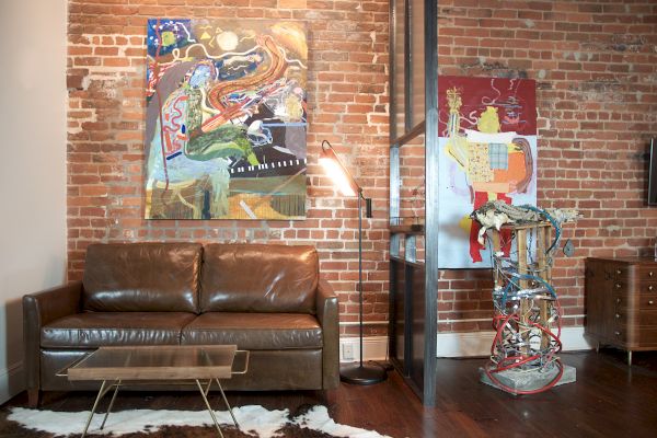 A living room with brick walls, a brown leather couch, vibrant abstract art, a unique lamp, and an eclectic sculpture next to a small wooden cabinet.