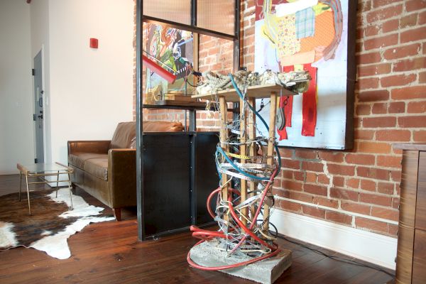 The image features a modern art installation with a chair, a coffee table, and a brick wall with paintings in the background.