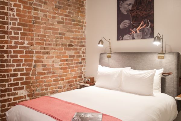 A cozy bedroom with a brick wall, a double bed with white linens, a red blanket, wall-mounted lamps, and artwork above the bed, and a book on the bed.