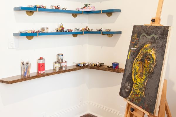 The image shows an art studio corner with blue shelves displaying small items, an easel with a painting, and art supplies on a wooden shelf.