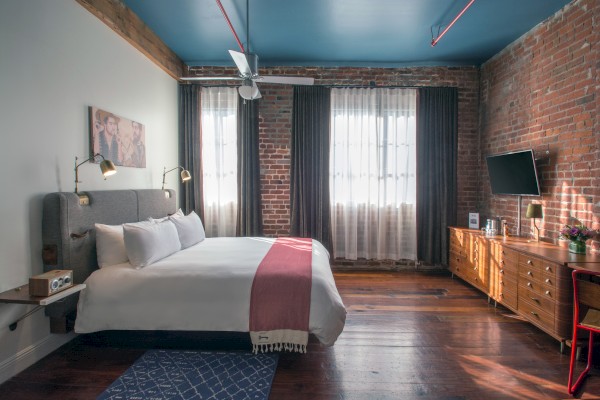 The image shows a cozy bedroom with a bed, brick walls, a TV, and large windows with curtains, creating a stylish and comfortable atmosphere.