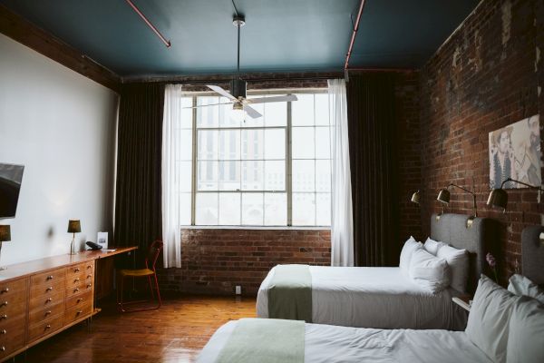 A cozy hotel room with two beds, a large window, wooden floors, a brick wall, a TV, a desk, and a ceiling fan, ending the sentence.
