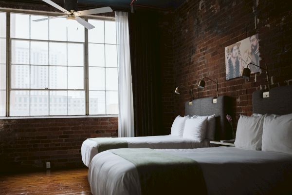 A cozy room with two beds, a large window, brick walls, a ceiling fan, and wall-mounted lights above each bed, and artwork on the wall.