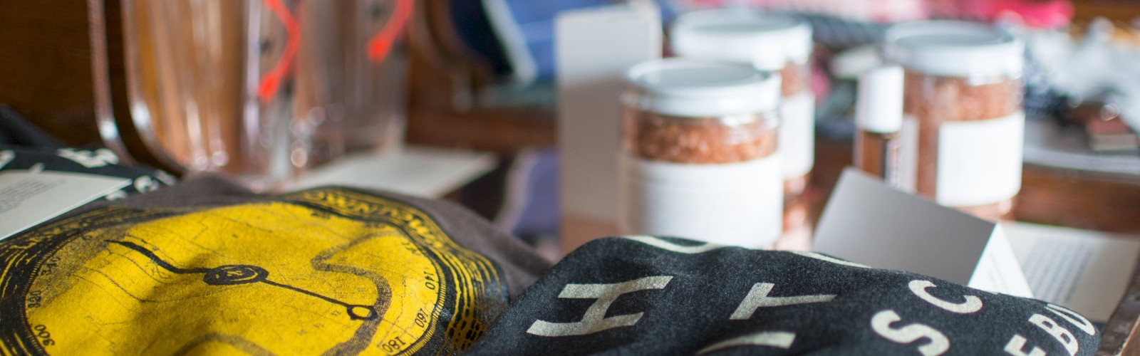 The image shows various merchandise on display, including T-shirts, glass cups, jars, and folded clothing items arranged on wooden surfaces.