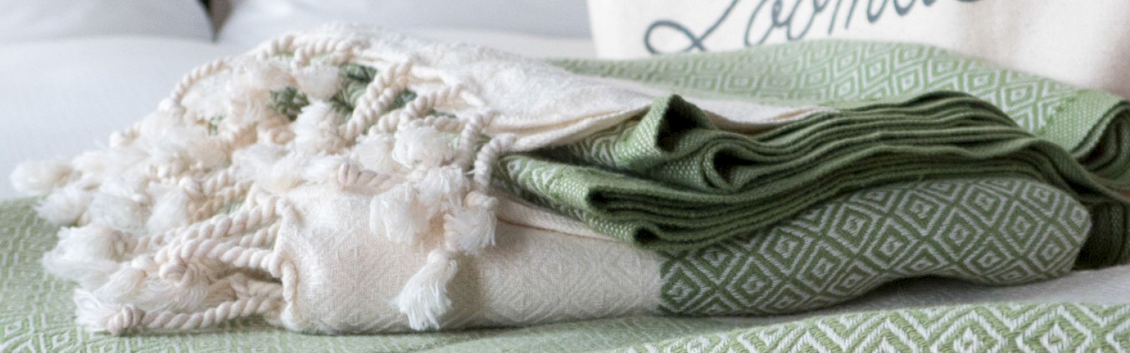 A folded green and white patterned blanket is topped with a cream tasseled throw, set on a bed with a white pillow and a cushion that says 