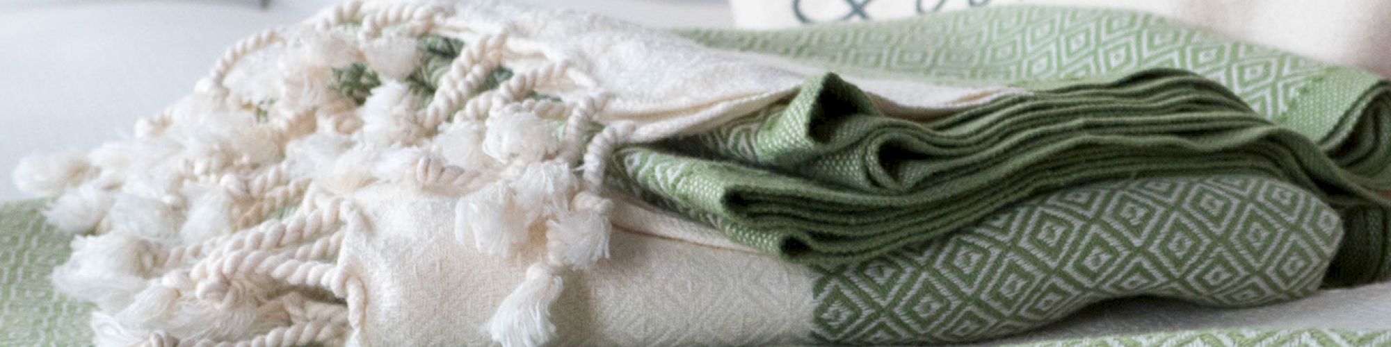 A folded green and white patterned blanket is topped with a cream tasseled throw, set on a bed with a white pillow and a cushion that says 