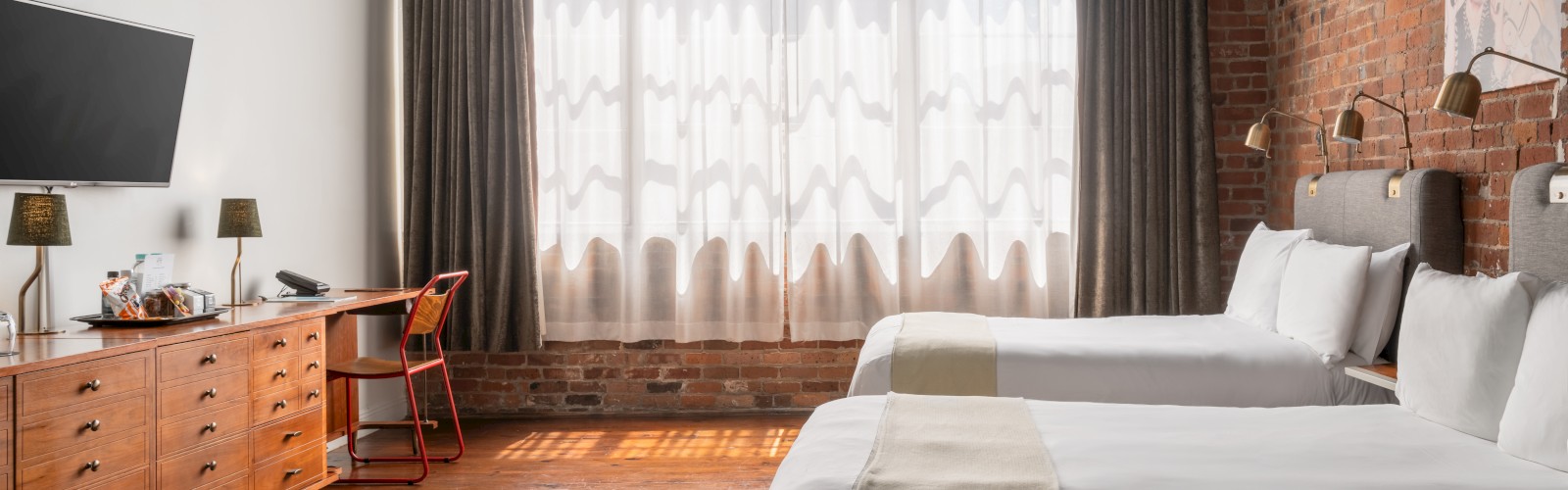 The image shows a brightly lit hotel room with two beds, a mounted TV, large windows with sheer curtains, and a ceiling fan.