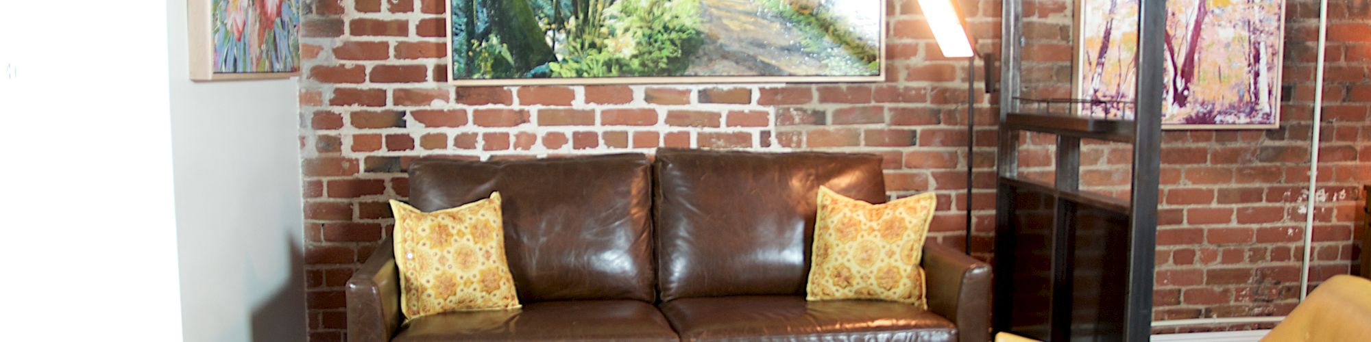 A cozy living room with a brown leather sofa, two yellow chairs, a coffee table, and nature-themed wall art on exposed brick walls.