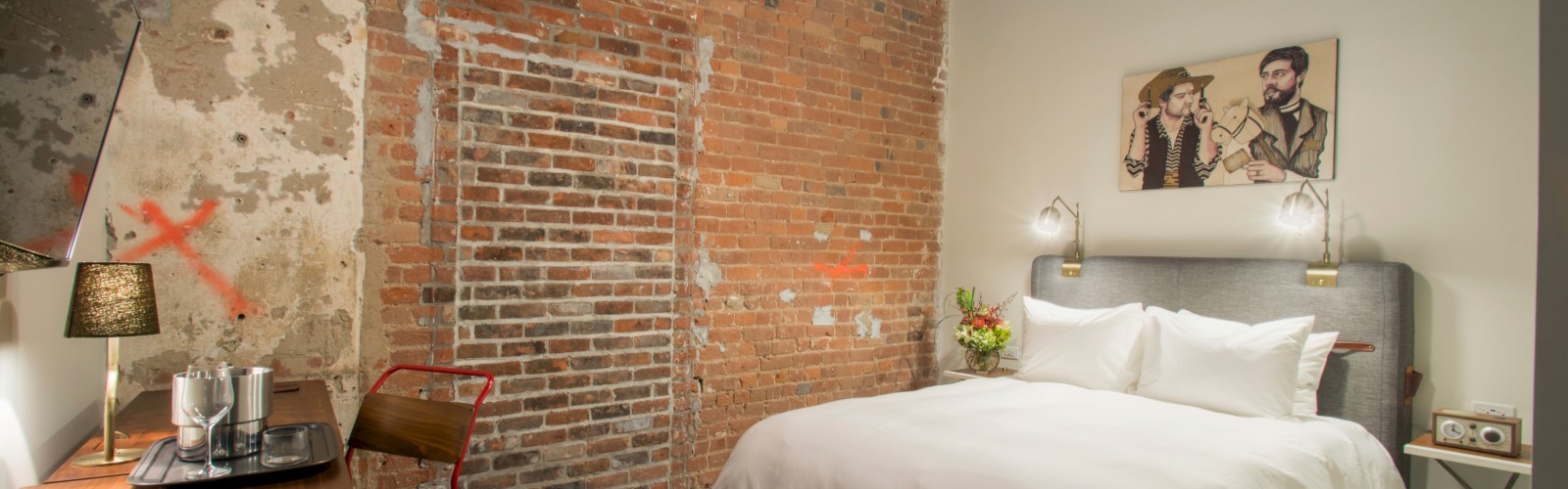 A cozy bedroom with exposed brick walls, a double bed, nightstands, a ceiling fan, and a small desk with a chair. A painting hangs above the bed.