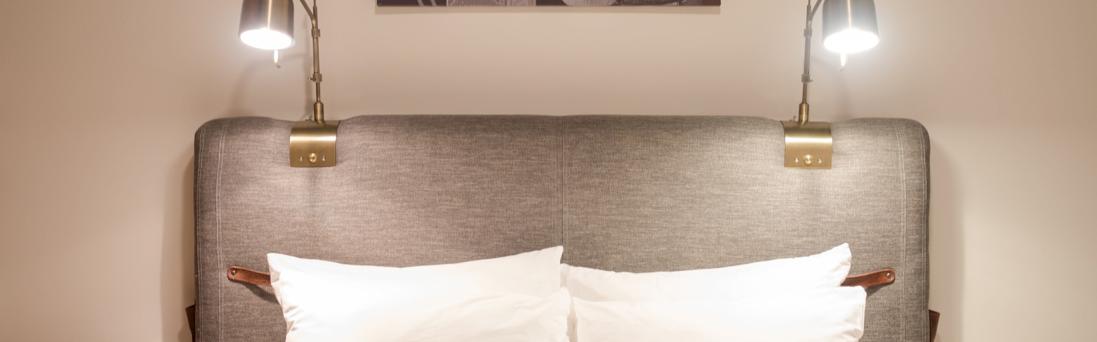 A neatly made bed with two white pillows, wall lamps on each side, nightstands with items, and a framed picture above the headboard ending the sentence.