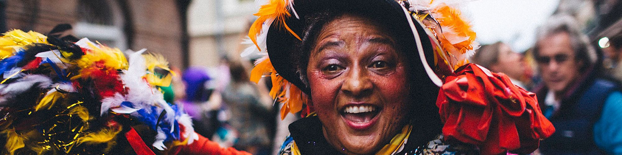 A joyful person in colorful attire and a festive hat surrounded by others, likely at a carnival or parade, smiles brightly at the camera.
