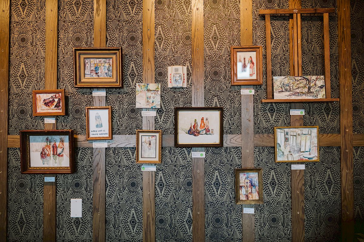 The image shows a wall adorned with various framed paintings of bottles and abstract designs, displayed on patterned wallpaper.