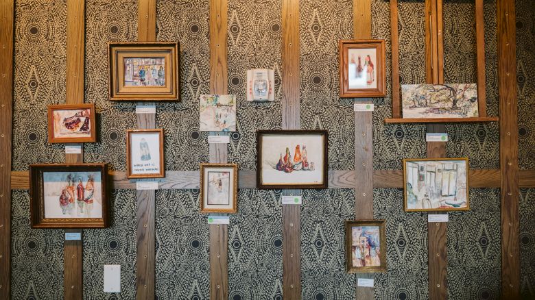 The image shows a wall adorned with various framed paintings of bottles and abstract designs, displayed on patterned wallpaper.