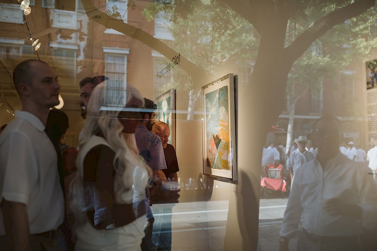 People are inside an art gallery, viewing paintings on the wall. There is a reflection on the glass showing the street and more people outside.