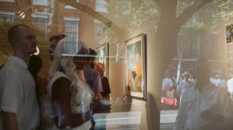 People are inside an art gallery, viewing paintings on the wall. There is a reflection on the glass showing the street and more people outside.