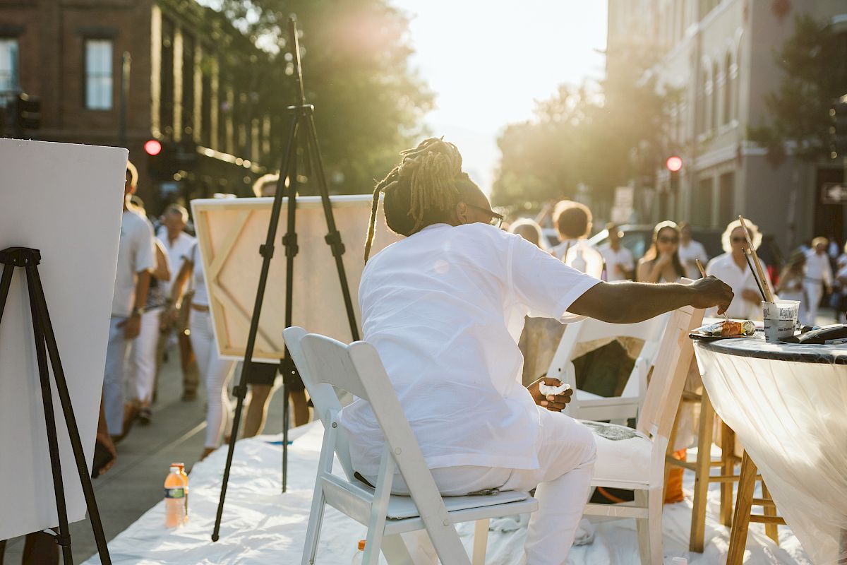 A person in white sits at an easel, engaging in painting outdoors amid a crowd in a sunlit urban setting.