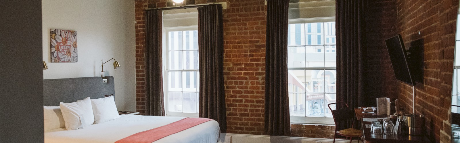 The image shows a cozy bedroom with a modern design featuring a large bed, brick walls, wooden flooring, a TV, and windows with curtains.