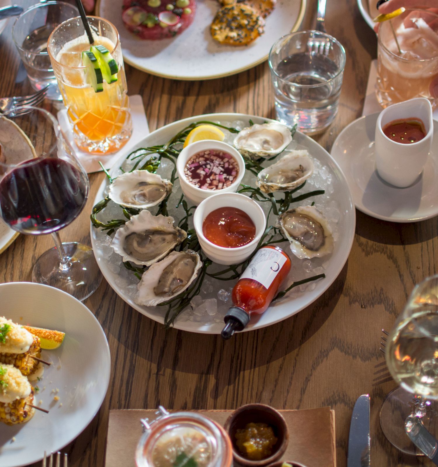A variety of dishes including oysters, drinks, and appetizers on a wooden table with multiple dining items, sauces, and hands reaching in.