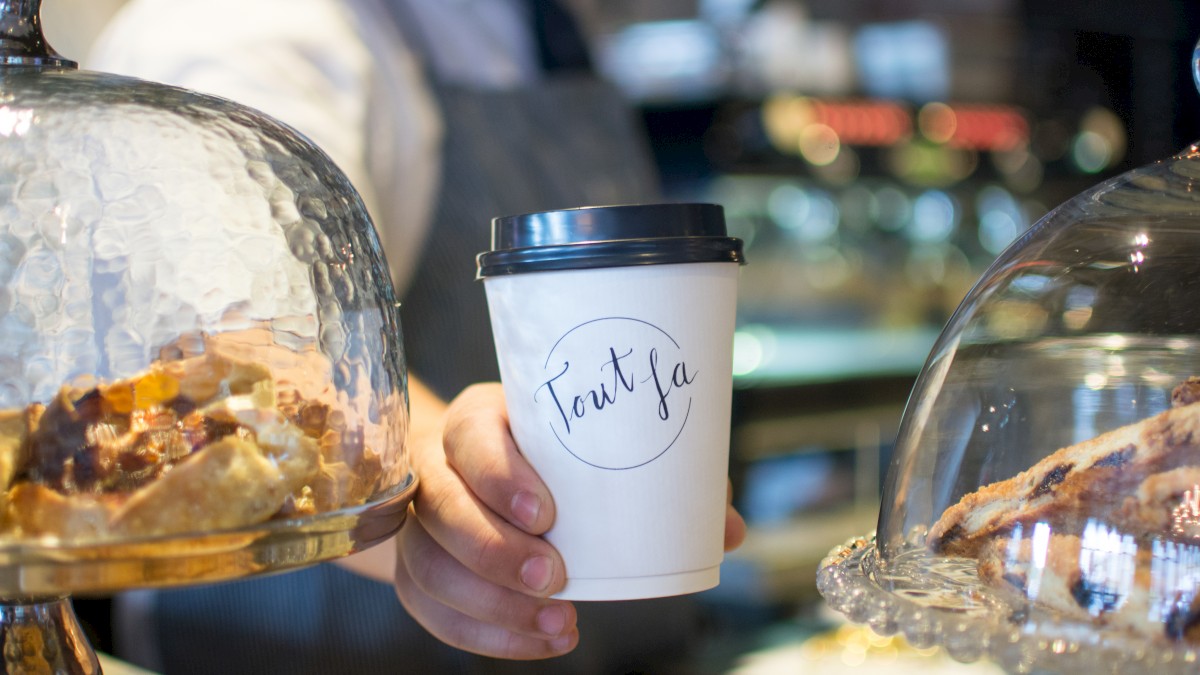 A person in a café holding a takeout coffee cup, surrounded by pastries under glass domes with blurred background.