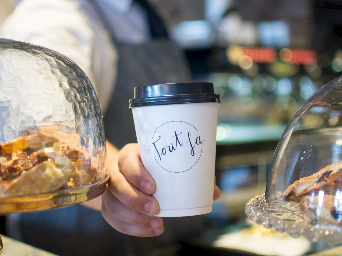 A person in a café holding a takeout coffee cup, surrounded by pastries under glass domes with blurred background.