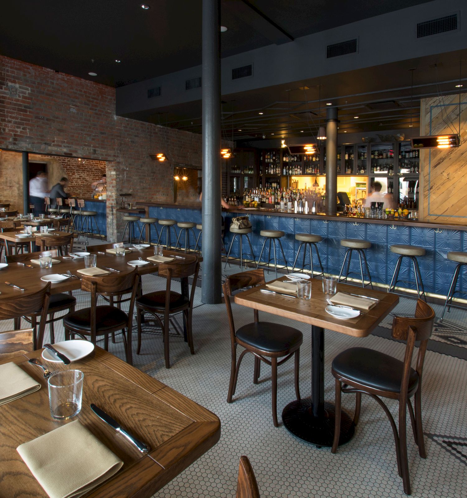 A modern restaurant with wooden tables set with plates and cutlery, brick walls, a blue-tiled bar with high stools, and a cozy ambiance.