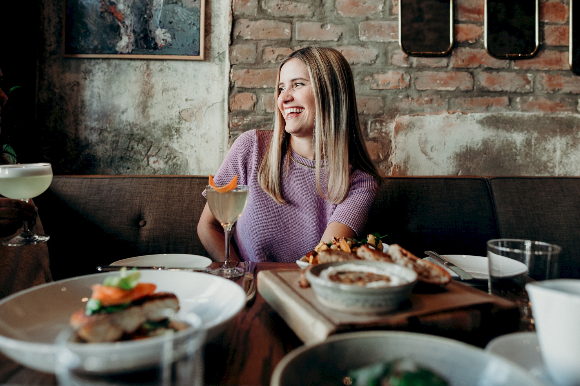 A person in a purple shirt is sitting at a restaurant table, smiling, with various dishes and a cocktail around them.