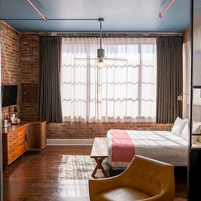 A cozy room with a bed, dresser, curtains, chair, and artwork, featuring exposed brick walls, wooden floors, and ample natural light.