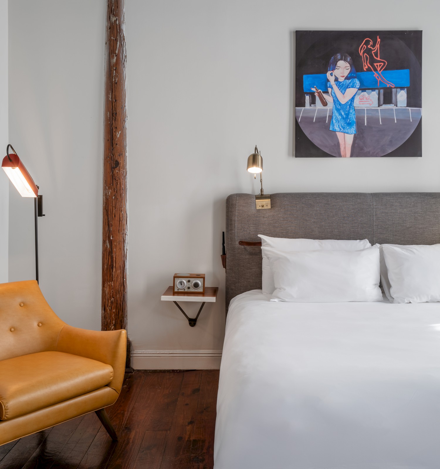 A cozy bedroom with a large bed, wall-mounted lamps, a modern chair, and a painting above the bed. The room has a minimalist aesthetic.