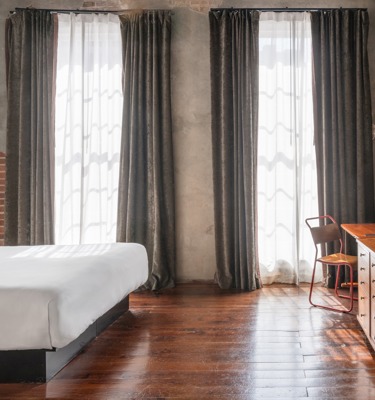 A minimalist bedroom with a large bed, dark curtains, wooden flooring, wall-mounted TV, and a rustic desk with a chair by two large windows.