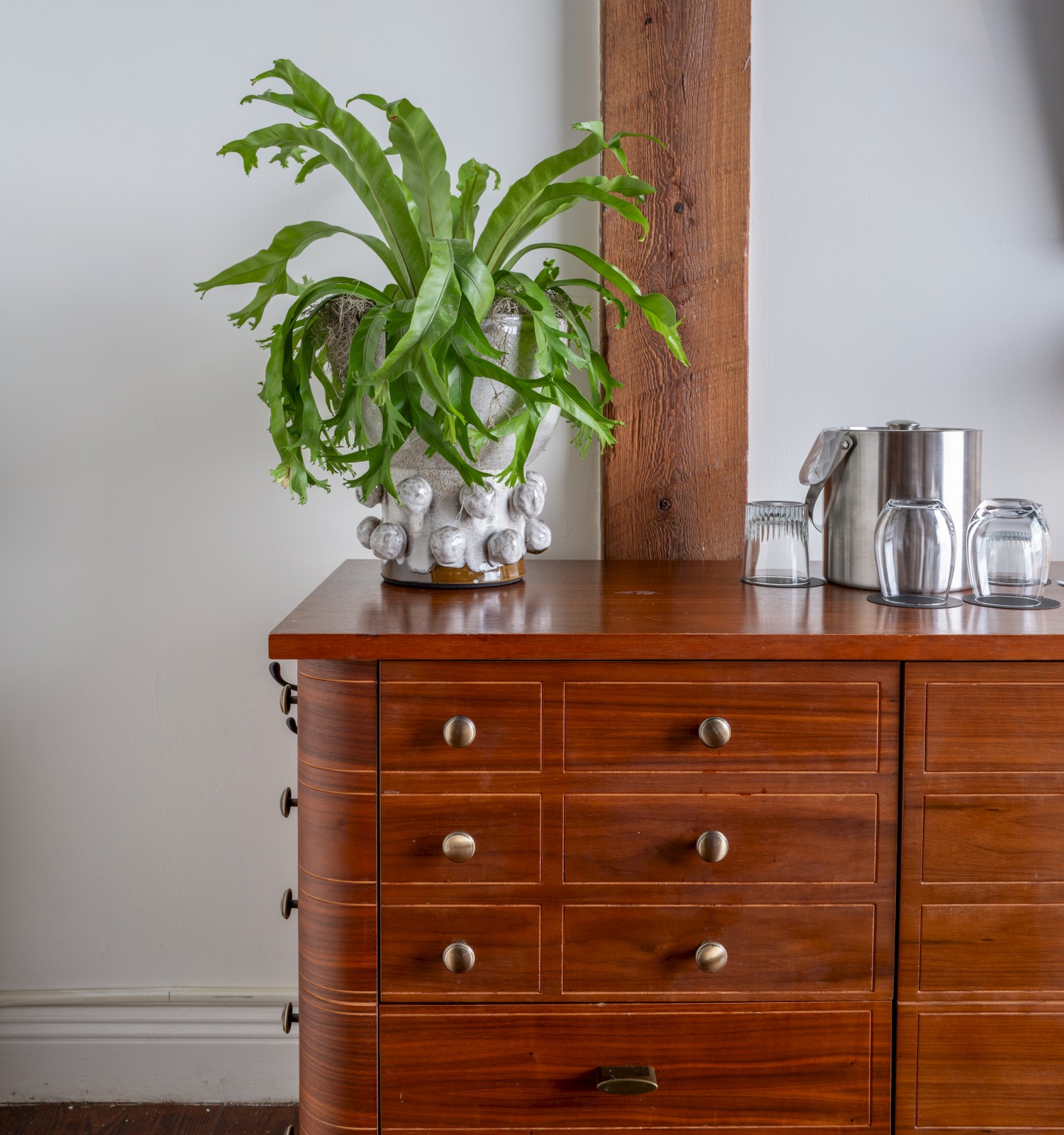 A wooden sideboard with drawers, a potted plant, glasses, and a metal pitcher stand against a white wall and wooden beam.