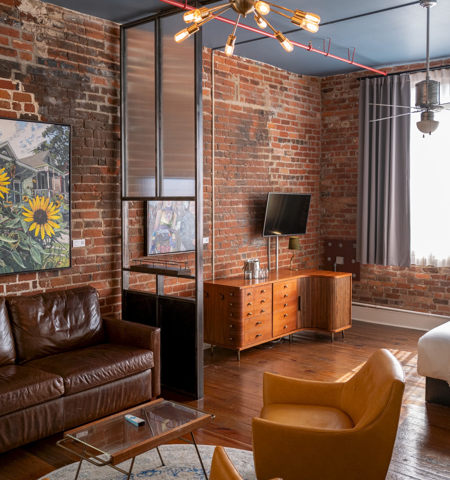 A loft-style room with exposed brick walls, a leather sofa, armchair, TV, and wall art featuring sunflowers. A bed is near a large window.