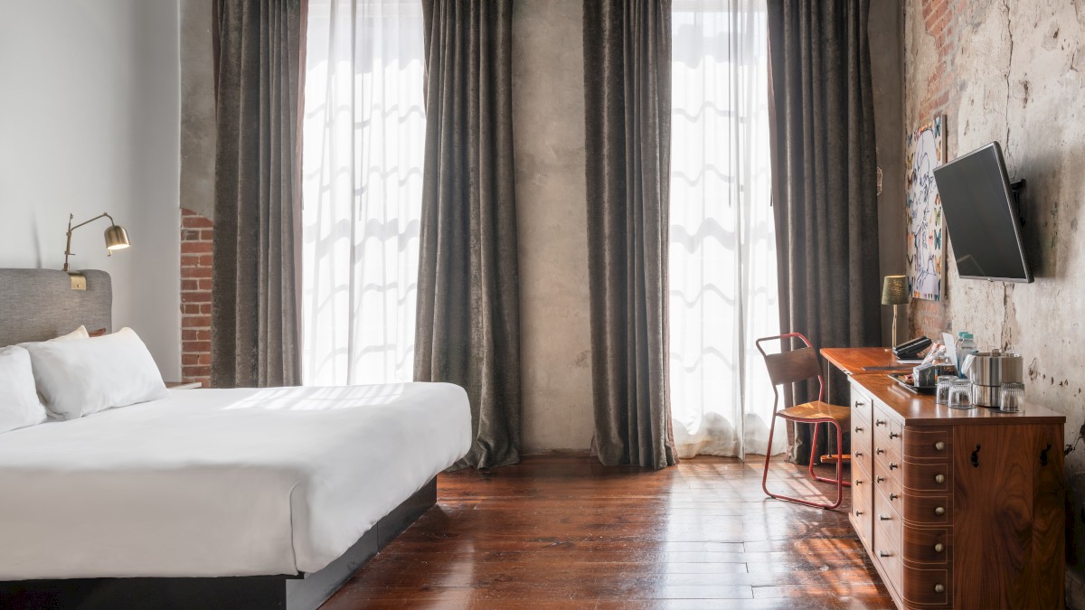 A stylish bedroom with a large bed, dark curtains, a flat-screen TV, wooden floors, and a desk with a chair by the window.